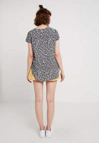 Černý top s krátkými rukávy a květinovým vzorem, vyrobený z lehké látky, s kulatým zakončením, kombinovaný se žlutou sukní a bílými botami.