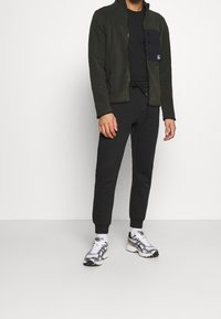 Fleece jacket in dark green with a zippered front, black chest pocket, paired with black joggers and silver athletic shoes.