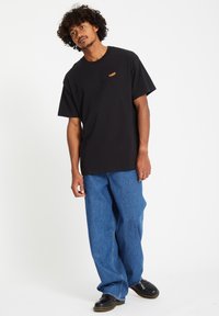 Zwarte katoenen t-shirt met korte mouwen en een oranje logo op de linkerkant van de borst, gecombineerd met losse blauwe denim broeken en zwarte schoenen.