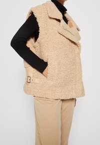 Ärmellose beige Sherpa-Weste mit Reverskragen und Schnallen, getragen über einem schwarzen gerippten Rollkragenpullover und beigen Hosen.