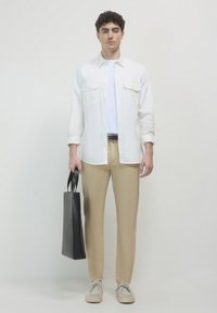 White button-up shirt over a white t-shirt, beige trousers, gray sneakers, and holding a black tote bag. Smooth fabric and tailored fit.