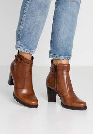 Bottes marron en cuir à talon montant jusqu'à la cheville avec fermeture éclair latérale et boucle, portées avec un jean bleu clair retroussé sur un sol blanc.