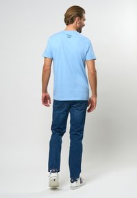 Svijetloplavi majica s kratkim rukavima i okruglim izrezom, s logom na leđima, u kombinaciji s tamnoplavim trapericama i bijelim tenisicama.