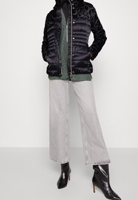 Veste rembourrée noire avec un empiècement en velours à imprimé léopard, associée à un pull en maille texturée verte, un jean à jambes larges gris clair, et des bottines à talons noires.