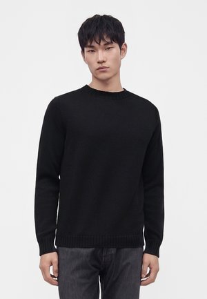 Jeune homme aux cheveux courts noirs portant un pull noir uni et un pantalon sombre, debout devant un fond clair uni.