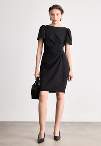 Melna kleita ar pieguļošu siluetu, īsām pūkām un drapētu priekšpusi. Saskaņota ar melniem augstpapēžu apaviem ar norādītu purngalu un mazu teksturētu rokassomu.