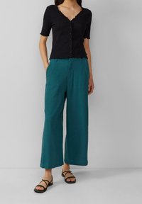 Black, short-sleeved, V-neck top with scalloped edges; paired with teal wide-leg pants. Black strappy sandals complete the outfit.
