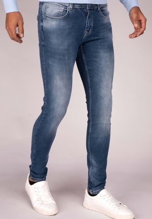 Homme portant un jean slim bleu denim et des baskets blanches, debout, les mains détendues le long du corps, sur fond neutre.