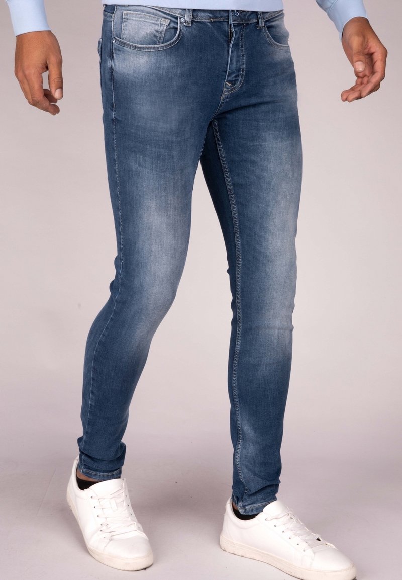 Homme portant un jean slim bleu denim et des baskets blanches, debout, les mains détendues le long du corps, sur fond neutre.