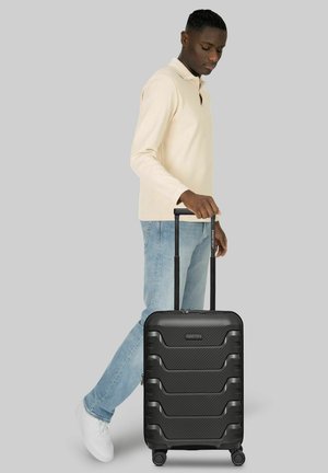 Mann in beige langärmligem Hemd und Jeans zieht einen schwarzen Hartschalenkoffer mit ausgezogenem Griff auf vier Rädern.