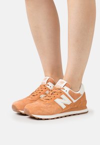 Osoba w pomarańczowych zamszowych sneakersach New Balance 574 z białymi akcentami, stojąca na neutralnym tle.