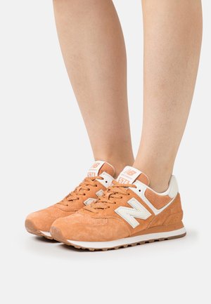 Osoba v oranžových semišových teniskách New Balance 574 s bílými akcenty, stojící na neutrálním pozadí.