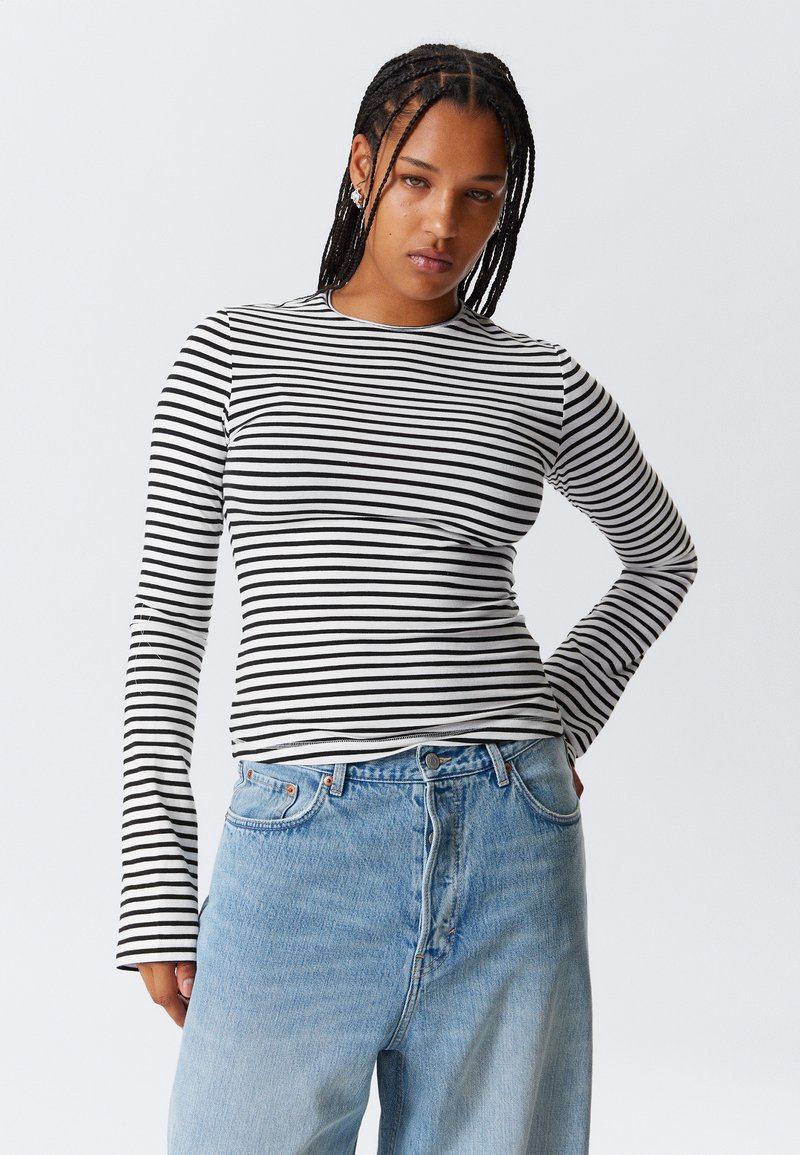 Femme avec des cheveux tressés portant un top à rayures noires et blanches à manches longues et un jean taille haute bleu clair, posant avec une main sur la hanche.