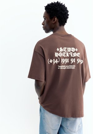 Jeune homme aux cheveux courts et bouclés portant un t-shirt marron oversize avec un texte graphique blanc dans le dos et un jean bleu clair.