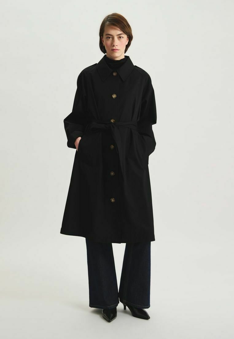 Manteau noir en trench avec un grand col, fermeture par boutons à l'avant et ceinture. Fabriqué en tissu lisse, il présente deux poches latérales et a une coupe décontractée.