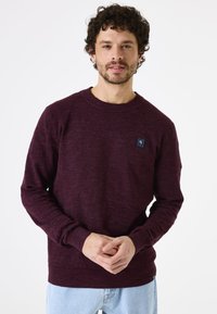 Bordeaux brei sweater met een ronde halslijn, geribde manchetten en zoom. Voorzien van een klein blauw logo-patch op de borst. Klassieke pasvorm.
