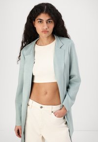 Světlomodrý dlouhý blazer s hladkou texturou, kombinovaný s upnutým krémovým cropped topem a bílými kalhotami s vysokým pasem. Vyznačuje se čistými liniemi a přiléhavým střihem.