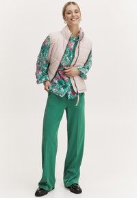 Vesta captușă roz deschis peste o bluză florală verde și pantaloni verzi largi. Pantofii negri completează ținuta, având un design fără cusături.