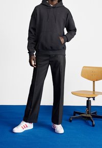 Černá mikina s kapucí a přední kapsou, v kombinaci s přizpůsobenými černými kalhotami a bílými teniskami s červenými pruhy. Modré pozadí na podlaze.