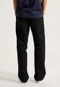 Black cotton pants with a loose fit, straight legs, and minimal design. Paired with white sneakers featuring red logo accents.