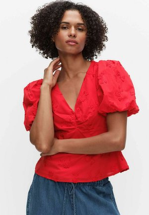 Vrouw met krullend haar, gekleed in een rode blouse met pofmouwen en bloemrijke borduursels, en blauwe denim broek met aantrekkoord, poseert met haar hand bij haar hals.