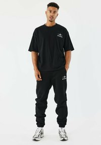 Camiseta negra de gran tamaño y joggers a juego hechos de algodón. La camiseta cuenta con un logo blanco; los joggers tienen un logo blanco en la cadera. Zapatillas con acentos en blanco y negro.
