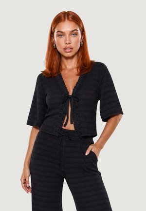 Femme aux cheveux roux portant un haut noir texturé à manches courtes noué à l'avant et un pantalon taille haute assorti avec un cordon.