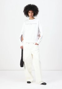 Felpa bianca oversize con maniche a taglio, con testo "TOMMY HILFIGER". Abbinata a pantaloni bianchi larghi e scarpe nere. Il modello tiene una borsa nera.