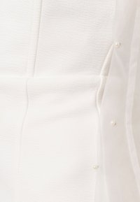 Tissu blanc texturé avec une superposition translucide ornée de petites épingles en perles sur le côté droit.