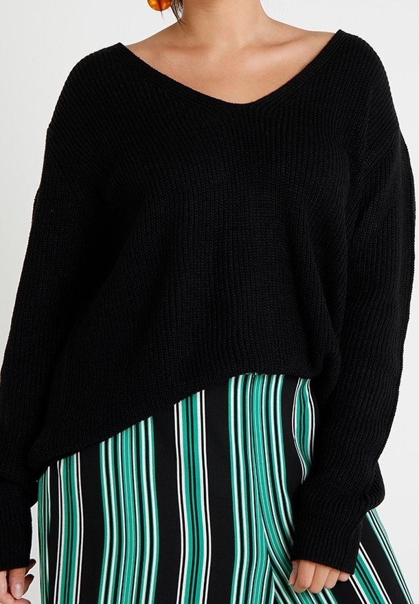 Femme portant un pull en tricot noir avec un col en V et une jupe rayée verticalement en vert, noir et blanc, sur un fond uni.