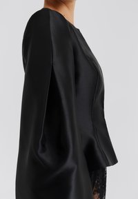 Μαύρο, δομημένο τοπ κατασκευασμένο από απαλό ύφασμα με στρογγυλή λαιμόκοψη και φουσκωτά μανίκια· διαθέτει διακριτική γυαλάδα και ραμμένη σχεδίαση.