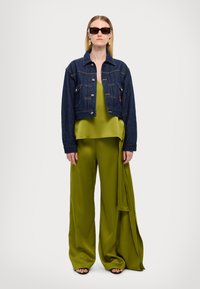 Jeansjakke med kort design, parret med en skinnende olivengrønn satengbluse og vide bukser med draperte detaljer. Svart solbriller brukt.