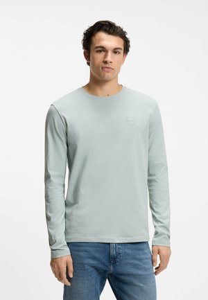 Giovane uomo con camicia a maniche lunghe grigio chiaro e jeans blu, in piedi contro uno sfondo bianco semplice, rivolto in avanti.