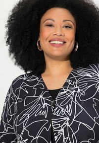 Cardigan noir avec un motif floral en ligne blanche. Il dispose d'un col en V, d'une fermeture à boutons et d'une texture de tissu lisse.