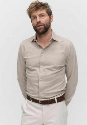 Beige Langarmhemd aus weichem Stoff mit klassischem Kragen, geknöpfter Vorderseite und subtiler Struktur, kombiniert mit weißen Hosen und einem braunen Gürtel.