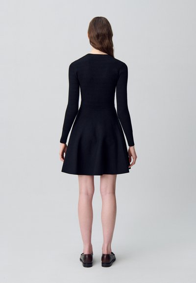 Donna in piedi di spalle che indossa un top fitted a maniche lunghe nero e una gonna svasata, con scarpe basse marrone scuro, su uno sfondo neutro.