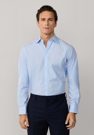 Homme aux cheveux courts bruns portant une chemise habillée bleu clair boutonnée et un pantalon bleu marine foncé, sur un fond uni.