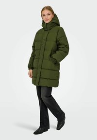 Zelená péřová bunda s vysokým límcem, předními knoflíky a bočními kapsami. Obsahuje prošívaný design a elastické manžety. Stylizováno s černými džíny.