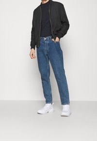 Schwarze Bomberjacke, navyblaues T-Shirt, hochgeschnittene blaue Jeans und weiße Sneaker. Glatter Stoff, lässige Passform und minimalistisches Design.
