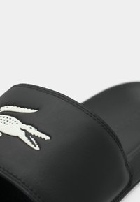 Sandale slide noire avec logo crocodile blanc sur la bride, texture lisse et coutures visibles autour des bords.