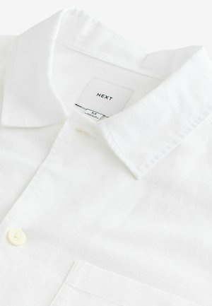 Chemise en lin blanche avec un col pointu, une fermeture à un bouton et une poche poitrine. Étiquette indiquant "NEXT," taille M. Texture douce.