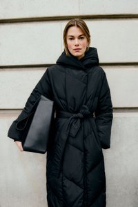Donna che indossa un lungo cappotto imbottito nero con cappuccio e cintura, mentre tiene una grande borsa a tracolla in pelle nera contro uno sfondo di muro di pietra.