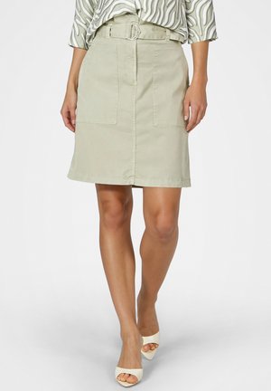 Frau trägt einen beigen knielangen Rock mit vorderen Taschen und Gürtel, kombiniert mit weißen offenen High Heels und gemustertem Oberteil.