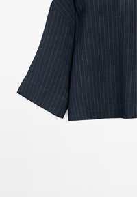 Tissu bleu foncé avec de fines rayures blanches verticales, montrant l'ourlet et la manche courte d'un vêtement sur un fond blanc.