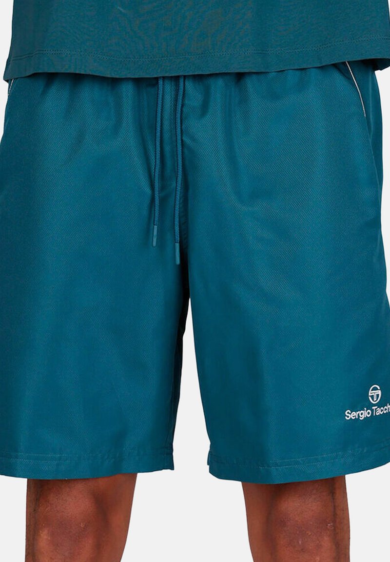 Shorts à cordon de serrage couleur sarcelle portés avec une chemise assortie, montrant les jambes de la mi-cuisse au genou, avec le logo Sergio Tacchini sur les shorts.