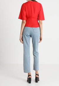 Kvinde iført en slim-fit rød bluse med flagrende ærmer, lyseblå højtaljede jeans og sorte blokhællede sandaler, stående med ryggen til.