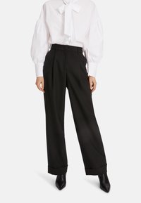 Schwarze weit geschnittene Hosen mit hoher Taille, ausgestattet mit einer gefalteten Vorderseite und umgeschlagenen Säumen, kombiniert mit einer weißen Bluse und schwarzen spitzen Schuhen.