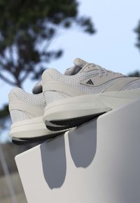 Baskets Adidas gris clair avec des détails en mesh et en daim reposant sur un rebord blanc en extérieur, montrant les semelles et les logos latéraux sur fond de ciel clair.