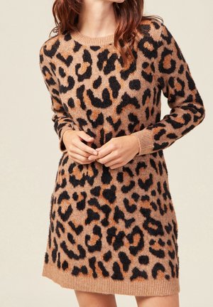 Femme portant une robe pull à manches longues, longueur genoux, avec un motif léopard marron et noir, les mains légèrement jointes devant elle.