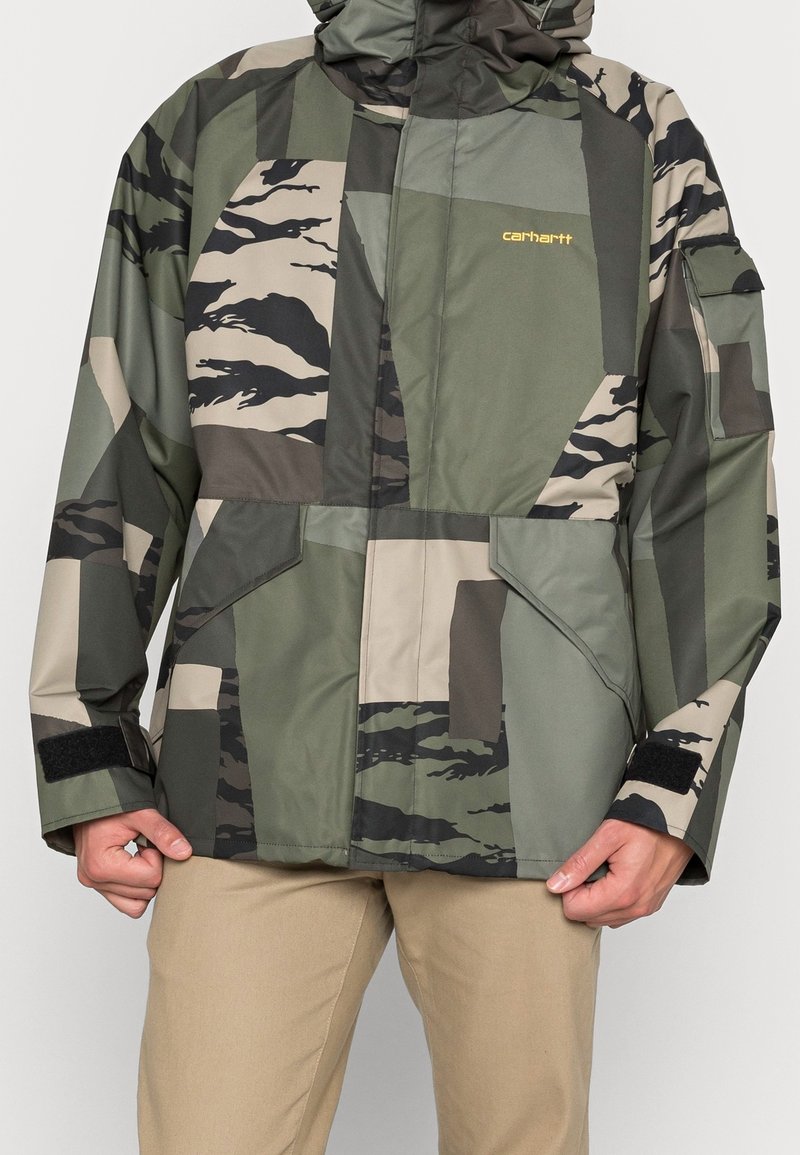 Homme portant une veste camouflage patchwork verte et beige avec capuche et pantalon beige, mains tenant l'ourlet de la veste.
